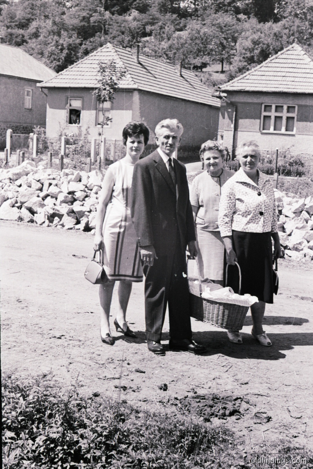 Family portrait from the 1960s or 70s, featuring a man in a suit, a young woman in a dress, and two older women carrying a picnic basket. Rural setting with modest homes and a stone pile in the background. Appears to be Eastern European. Charming snapshot of daily life.