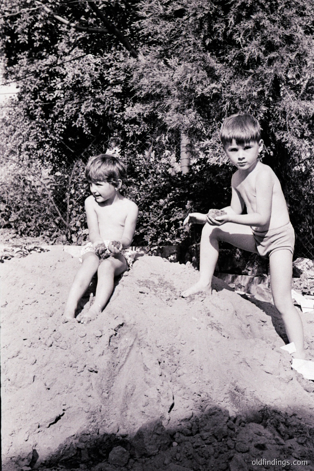 Two young boys sit atop a large sand dune. One is shirtless, holding handfuls of sand, while the other wears swim trunks and white sandals. Lush foliage forms a backdrop, suggesting a coastal location. Likely taken in the 1960s or 1970s, exhibiting classic documentary-style photography. Suitable for historical archives and design referencing childhood.