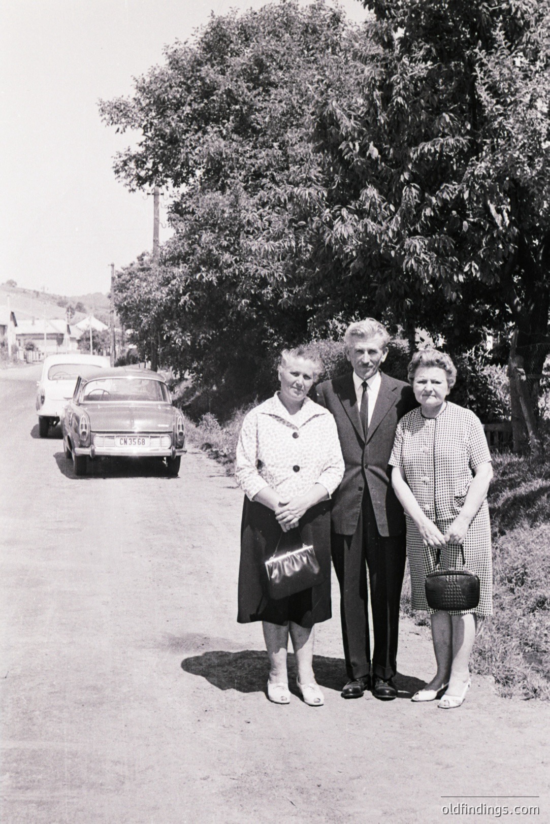 A posed black and white portrait depicts three formally dressed individuals standing on a rural road. A classic car is parked nearby. Likely 1960s, the subjects appear to be a family or group of friends. Clothing styles and vehicle suggest a mid-century setting. A glimpse of a building can be seen in the background.