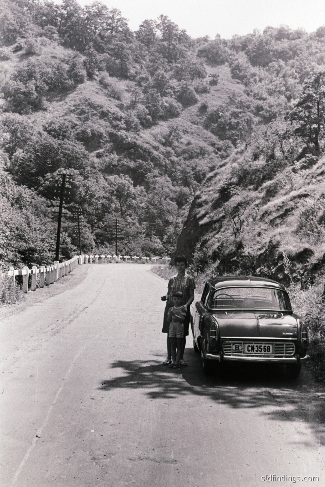 Monochrome image showing a woman and young child standing beside a vintage car on a winding mountain road. Dense, forested slopes rise on either side. Car’s plate reads “CN 35-68”. Likely 1960s or 70s, possibly Eastern European setting due to vehicle style. Illustrates travel & leisure, automotive design references.