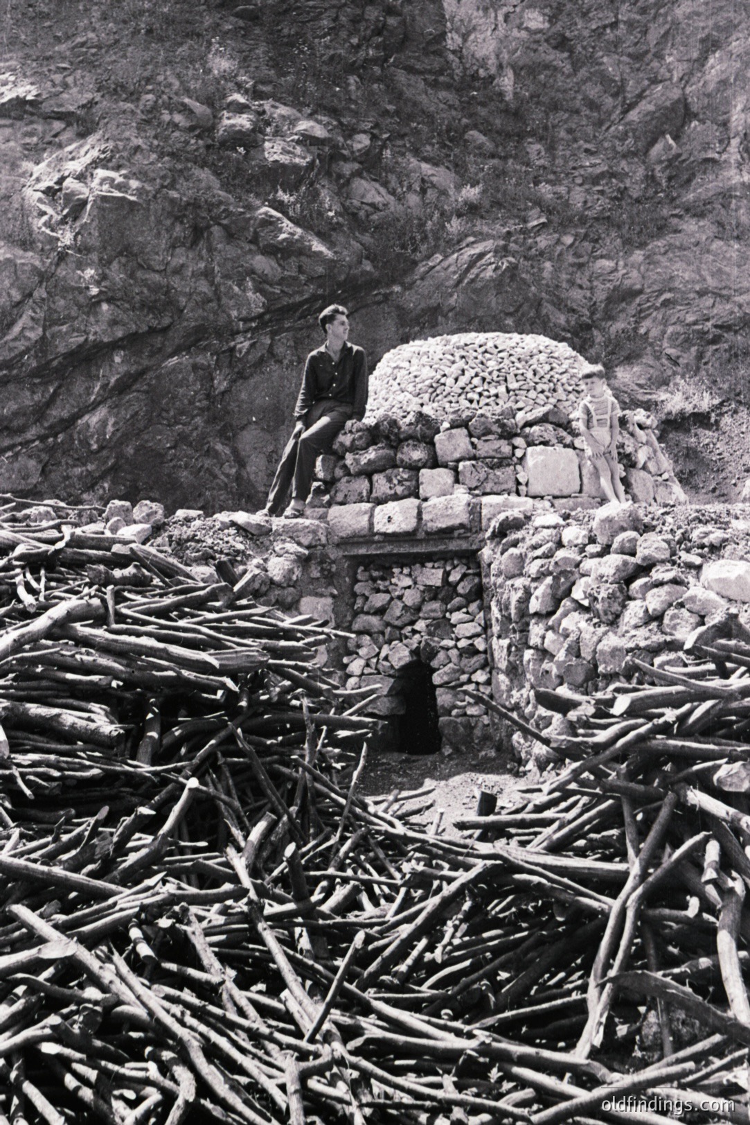 Man seated atop a rugged, dry-stone structure built into a rocky cliff face. The structure features a small opening and a domed roof. Debris of branches and driftwood litter the foreground. Appears to be a seaside or coastal location. Likely 1950s or 1960s documentary style.