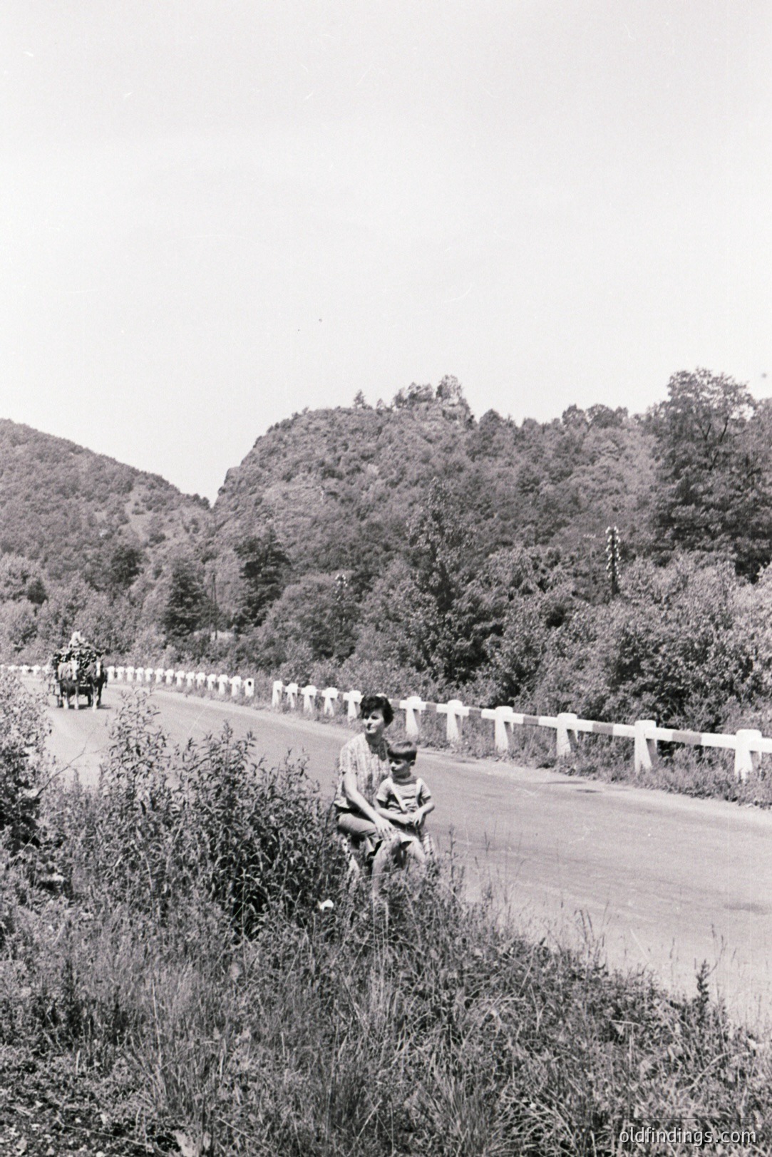 A woman holds a young child amidst roadside vegetation. Asphalt road bordered by a low stone wall leads toward a forested hillside. Likely a rural area, possibly Eastern Europe. Appears to be a snapshot from the 1960s or 70s. Documentary-style image, potential stock photo value.