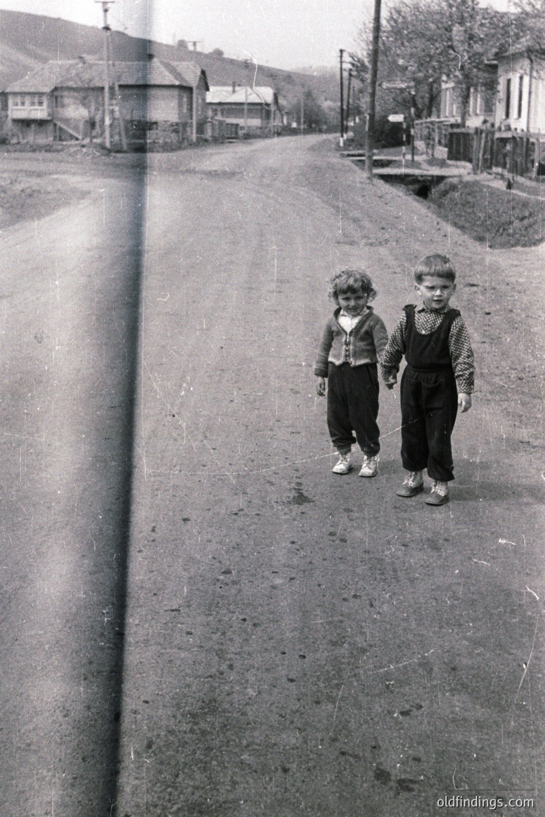 Two young boys, holding hands, walk down a dirt road lined with modest, older homes. Visible wear marks along the left edge suggest potential film damage. Likely rural setting, possibly Eastern Europe, mid-20th century. Clothing style indicates 1940s-1960s. Simple, evocative portrait.