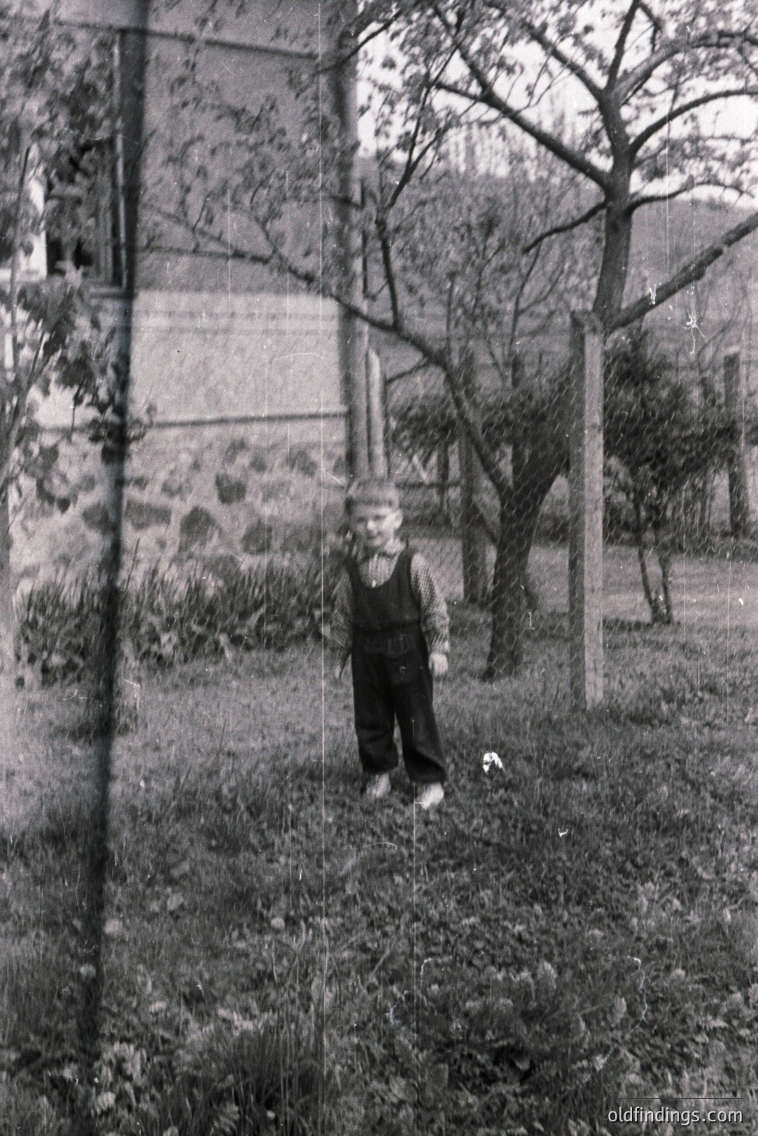 A young boy stands in a grassy yard, wearing denim overalls and a short-sleeved shirt. A stone building is visible in the background, partially obscured by foliage. The aged photograph exhibits signs of wear, including scratches. Likely a family snapshot from the mid-20th century.