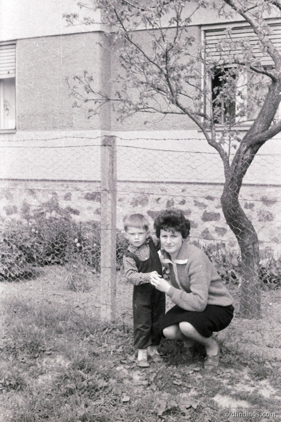A young boy in overalls stands alongside his mother, who kneels near a wire fence. A stark, modernist building forms the backdrop. Likely mid-20th century, perhaps 1960s, judging by clothing and architectural style. Captures a candid, family moment.