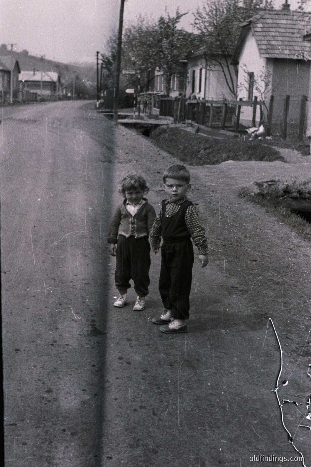 Two young boys stand on a rural road, dressed in traditional clothing - one in a patterned vest and dark trousers, the other in a collared shirt and similar pants. The scene suggests a 1960s or 70s Eastern European village setting, with simple homes and a natural landscape. The image displays signs of age and original processing.