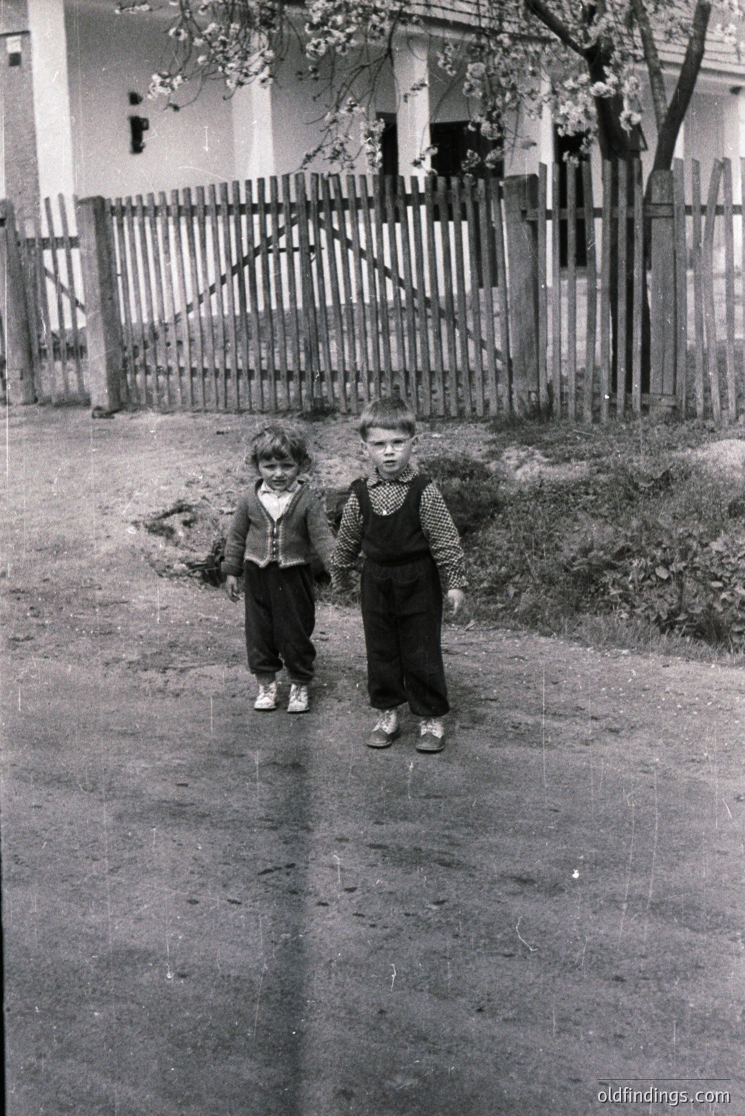Two young boys stand facing the camera on a wet paved road. They wear period-appropriate clothing; one in a corduroy jacket, the other in overalls and a collared shirt. A weathered wooden fence and older building with visible dormer window are in the background. Likely rural setting, possibly Eastern Europe, 1950s-1970s. Commercial value for family portraits, vintage style.