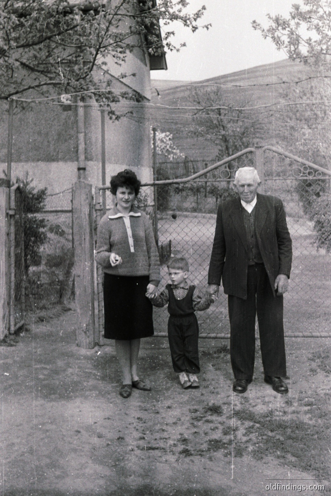 A family portrait: a woman, young child, and elderly man stand outside a modest home. The man and child hold hands. The hillside backdrop suggests an alpine or coastal location. Likely taken in the 1950s-1970s based on clothing styles. The scene evokes everyday life of the era.