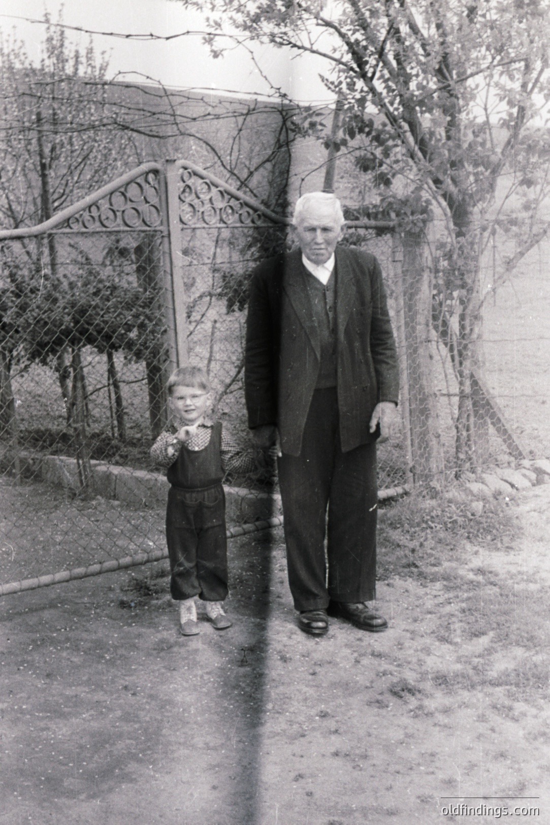 A young boy stands beside an older man, possibly a grandfather, outside a rustic home. The man wears a suit and tie; the boy, overalls. A decorative gate and overgrown foliage create a backdrop. Likely from the mid-20th century. A poignant, sentimental family portrait.