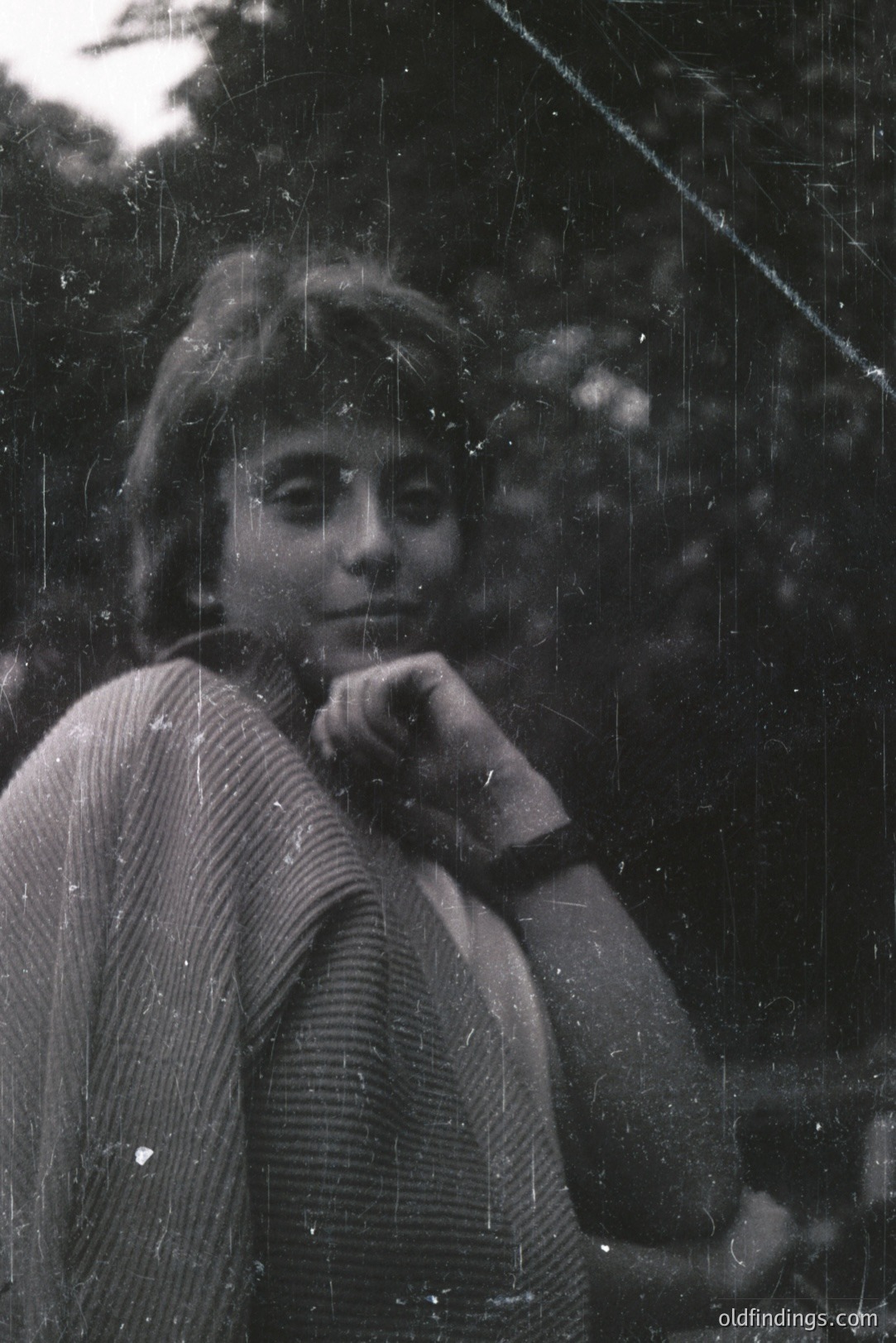 A young woman with short, layered hair poses outdoors, appearing relaxed. She wears a textured knit sweater and a dark wristwatch. Visible grain and scratches suggest a vintage film photograph, likely 1970s. Background foliage is softly blurred. A portrait study; potentially suitable for design reference.