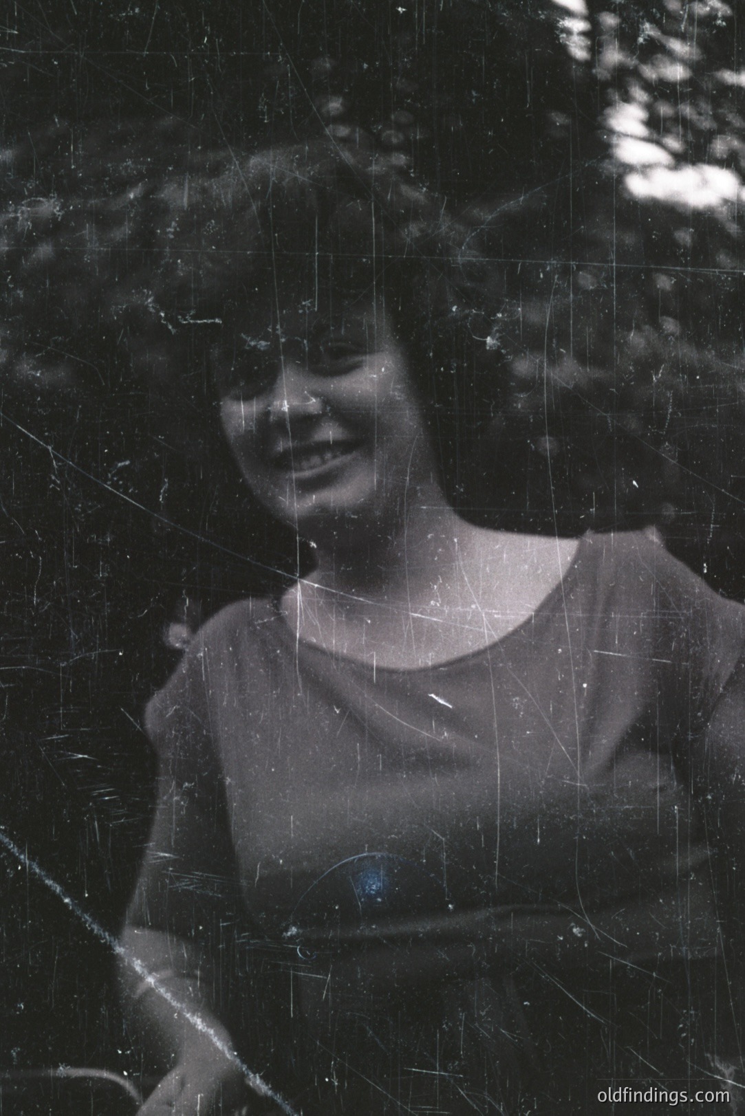 A black and white portrait of a young woman with wavy hair, smiling gently. She wears a simple, boat-neck top. The background features blurred foliage, suggesting an outdoor setting. Heavily scratched film indicates age and potential archival value. Likely 1970s or 1980s amateur photography.