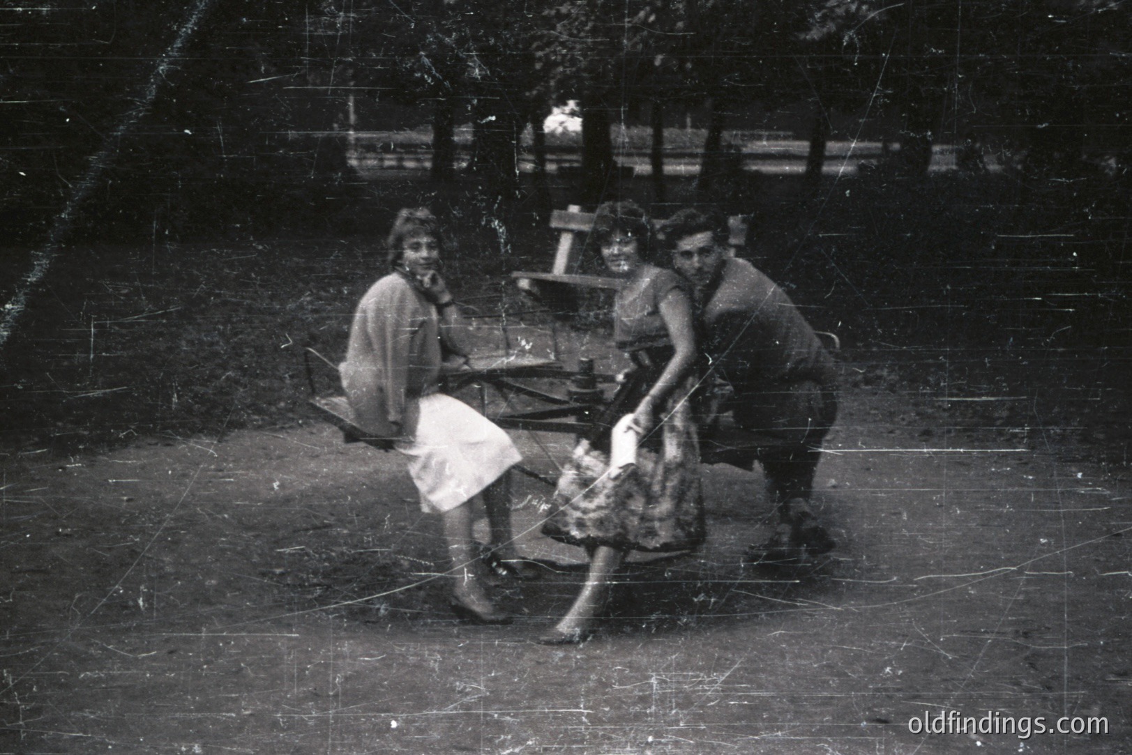 Three people seated on a picnic table in an outdoor setting. A woman in a dress, another in a skirt suit, and a man in a jacket are posed casually. Likely a family or group of friends. Appears to be a candid moment, circa 1960s. Potential for vintage lifestyle stock photography use.