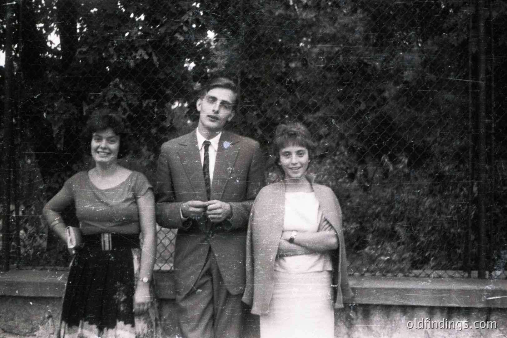 Three figures stand posed outdoors; a man in a suit and tie between two women in dresses. The setting appears to be a garden or park, with foliage visible behind them. Likely a formal portrait, c. 1960s-1970s. Captures a moment of social documentation.