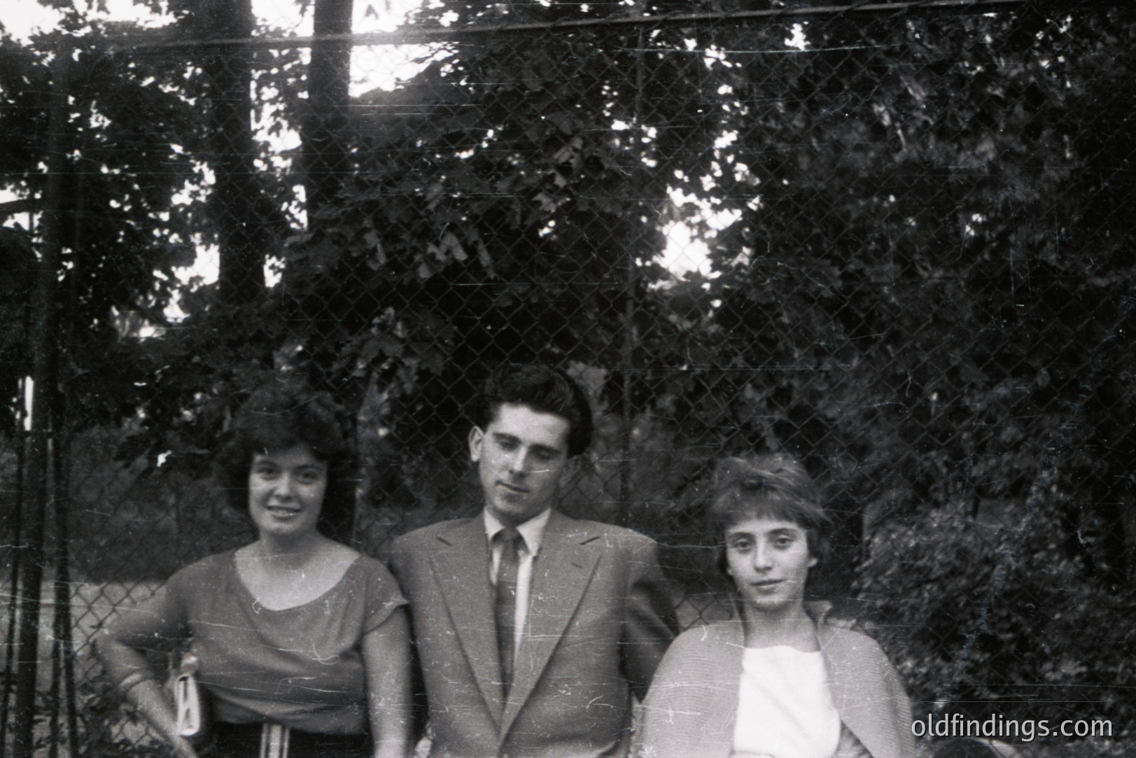 Three formally dressed individuals pose outdoors, possibly for a portrait. The man wears a suit and tie; the women sport 1960s-era hairstyles and attire. A glimpse of foliage and fencing suggests a garden or park setting. Photograph exhibits signs of age.