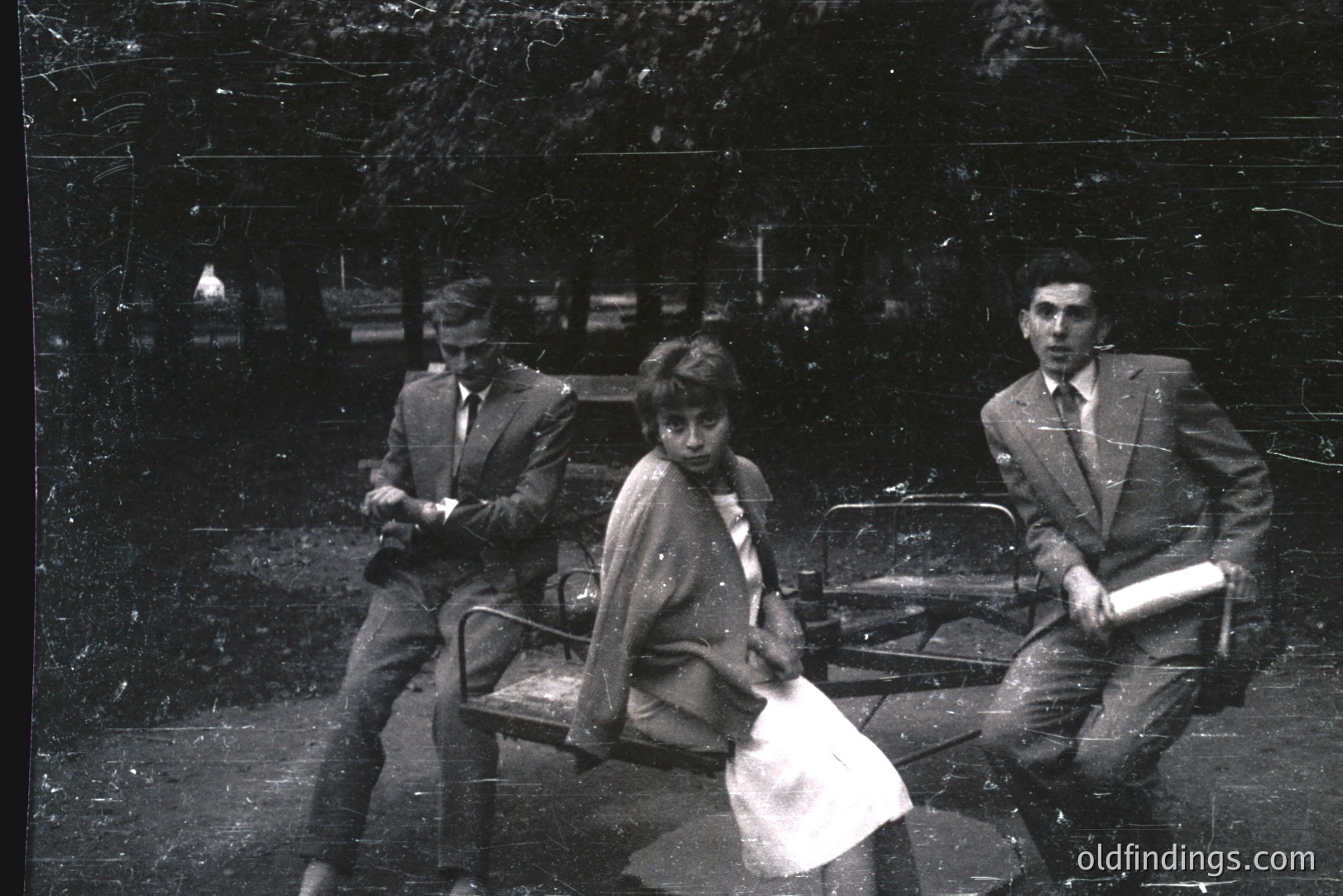Three figures posed on a circular bench in a park setting. Two young men in suits flank a young woman, who appears pensive. One man holds rolled papers. Likely a candid portrait from the 1960s or 70s, evocative of post-war European formality.