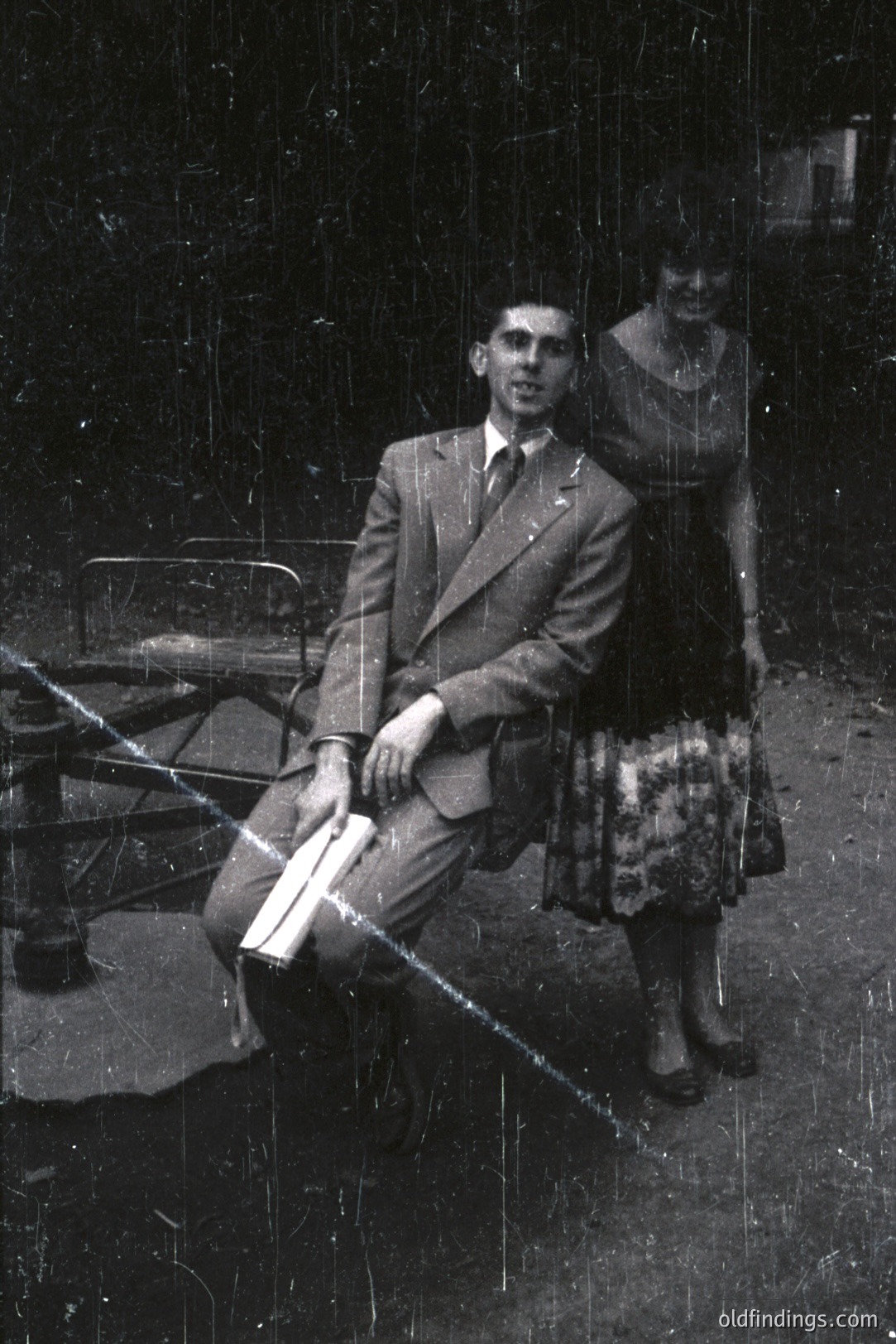 A formally attired man sits on a metal chair, holding sheet music. A woman in a floral dress stands beside him. Appears to be an outdoor studio portrait, likely 1960s or 1970s. Significant image degradation and scratches.