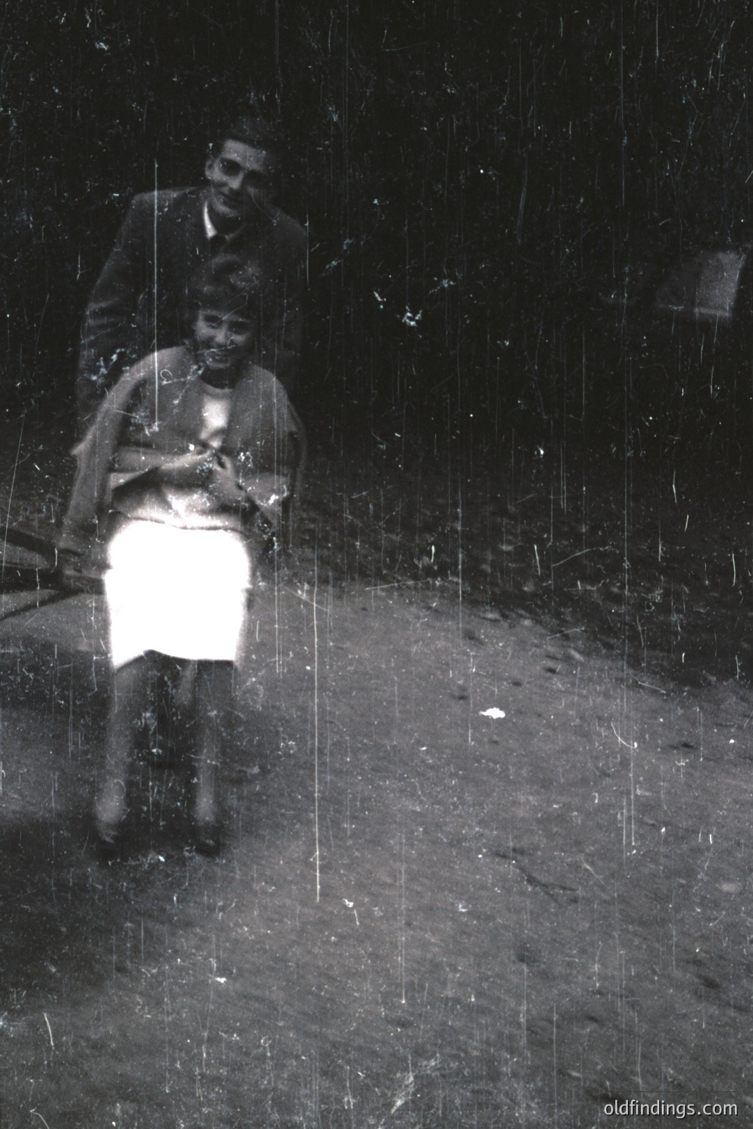 A seated woman in a tailored suit and a man in a jacket stand outdoors. The woman holds what appears to be a small book or prayer beads. The photo's texture reveals film grain and age. Likely a candid family portrait, c. 1950s - 1970s. Subject matter suitable for historical research or design.