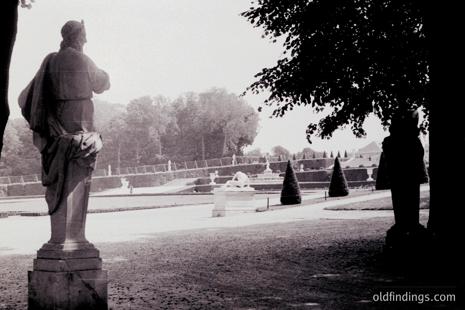 Classical statue overlooks manicured gardens with fountains and topiaries. Formal landscaping suggests a European estate, possibly French or Italian design. Likely a photograph from the mid-20th century, showcasing landscape architecture and statuary. A well-suited image for design or historical reference.