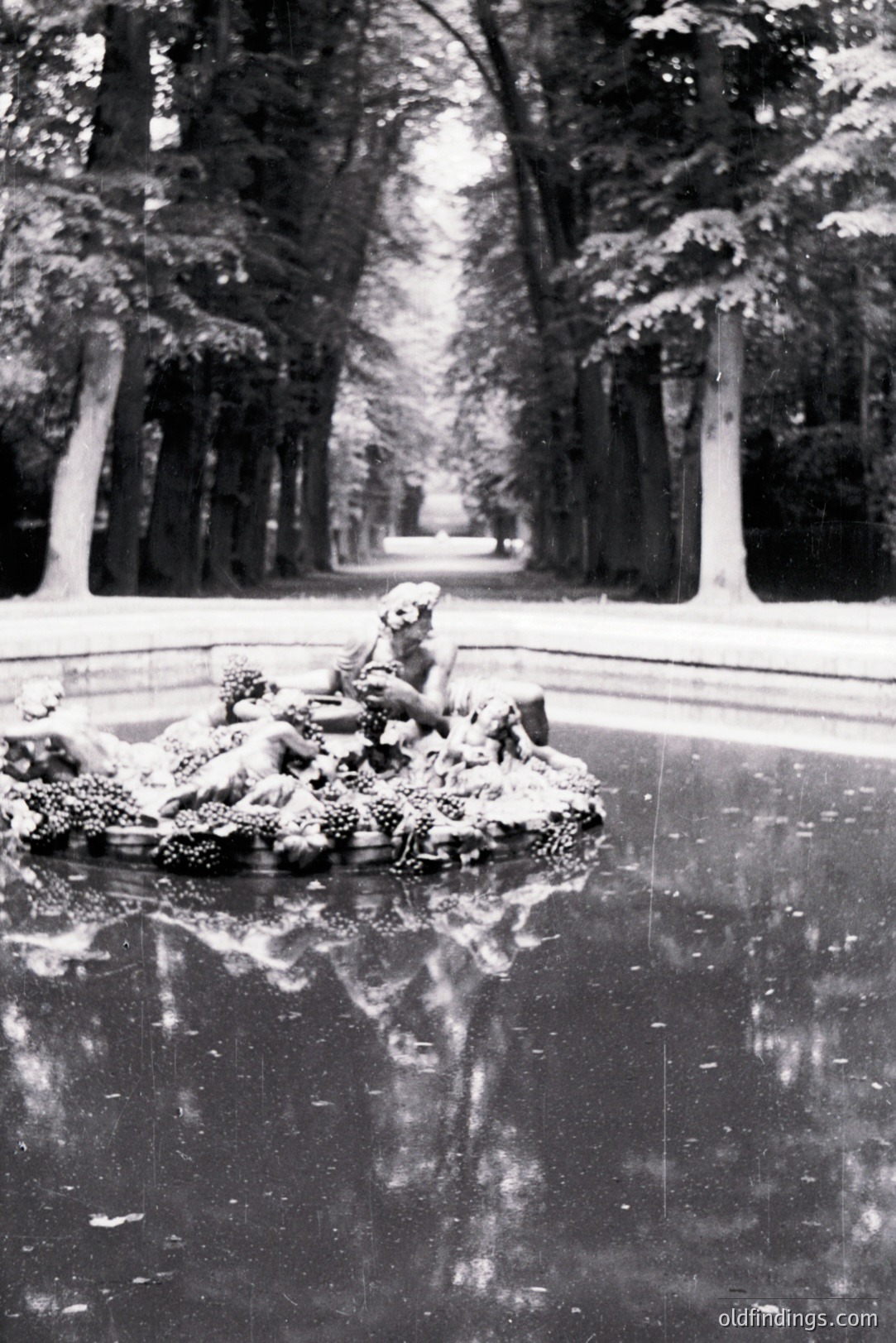 Ornate sculpture atop a dark pool, framed by a straight, tree-lined avenue. Statue depicts figures amidst foliage, reflected in still water. Likely an estate garden or formal park setting, possibly 20th century. A serene and formal composition.