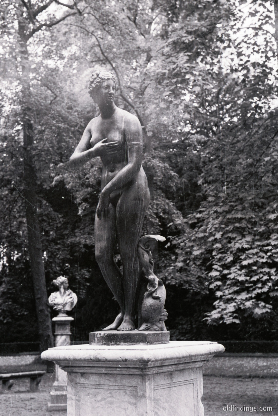 A bronze statue of a woman in a classical pose stands on a stone pedestal within a lush, leafy park setting. She appears to be gesturing towards herself, with a contemplative expression. A bust is visible on a secondary pedestal in the background. Likely 19th-century, possibly French or German.