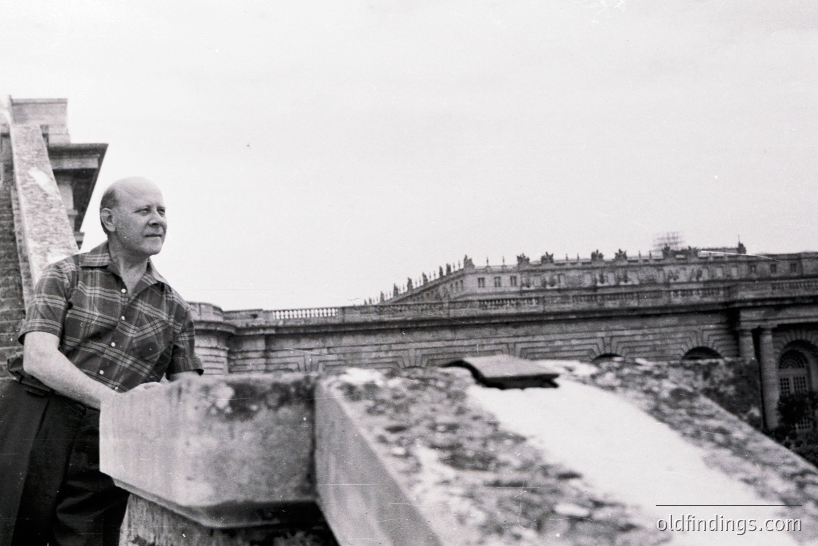 A man in a plaid shirt stands on a rooftop, leaning on a weathered stone parapet. Behind him stretches a monumental, ornate building with a long balustrade. Likely taken in the mid-20th century, the image evokes a sense of architectural grandeur. Possible location: Europe. Useful for architectural studies.