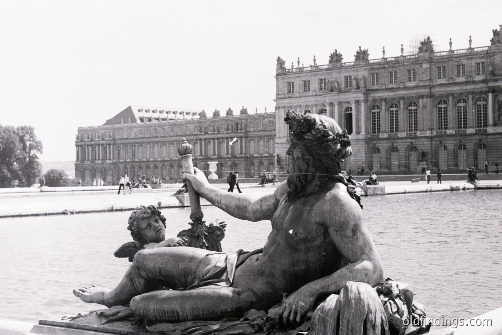 Statue of a reclining male figure, likely a deity, with a smaller cherubic figure nearby, dominates foreground. Behind, the grand facade of a palace—likely the Palace of Versailles—is visible across a reflecting pool. Likely 1960s/70s black & white photography. Ideal for architectural or art historical studies.