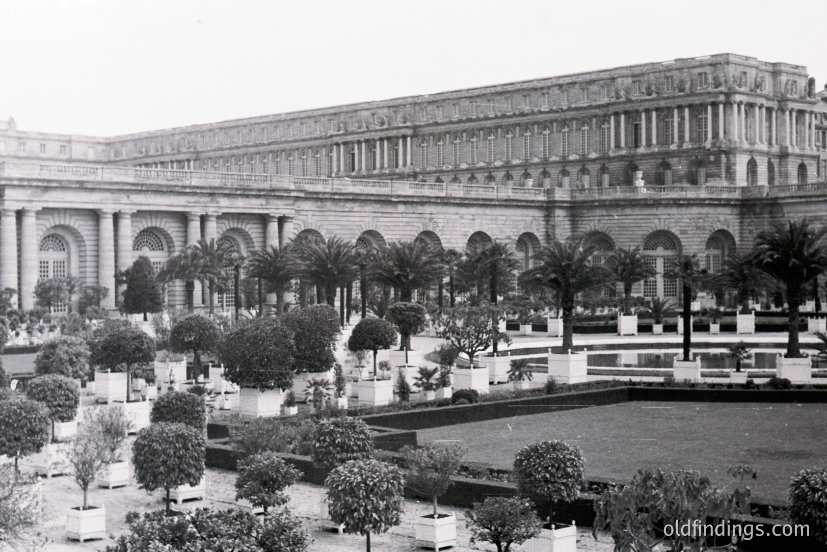 Formal garden with meticulously manicured boxwood shrubs and palms precedes a grand, classically-inspired building. The structure features a colonnaded arcade and layered, ornate detailing. Likely a palace or institutional building. Estimated date: early-mid 20th century. Potential architectural reference for design projects.