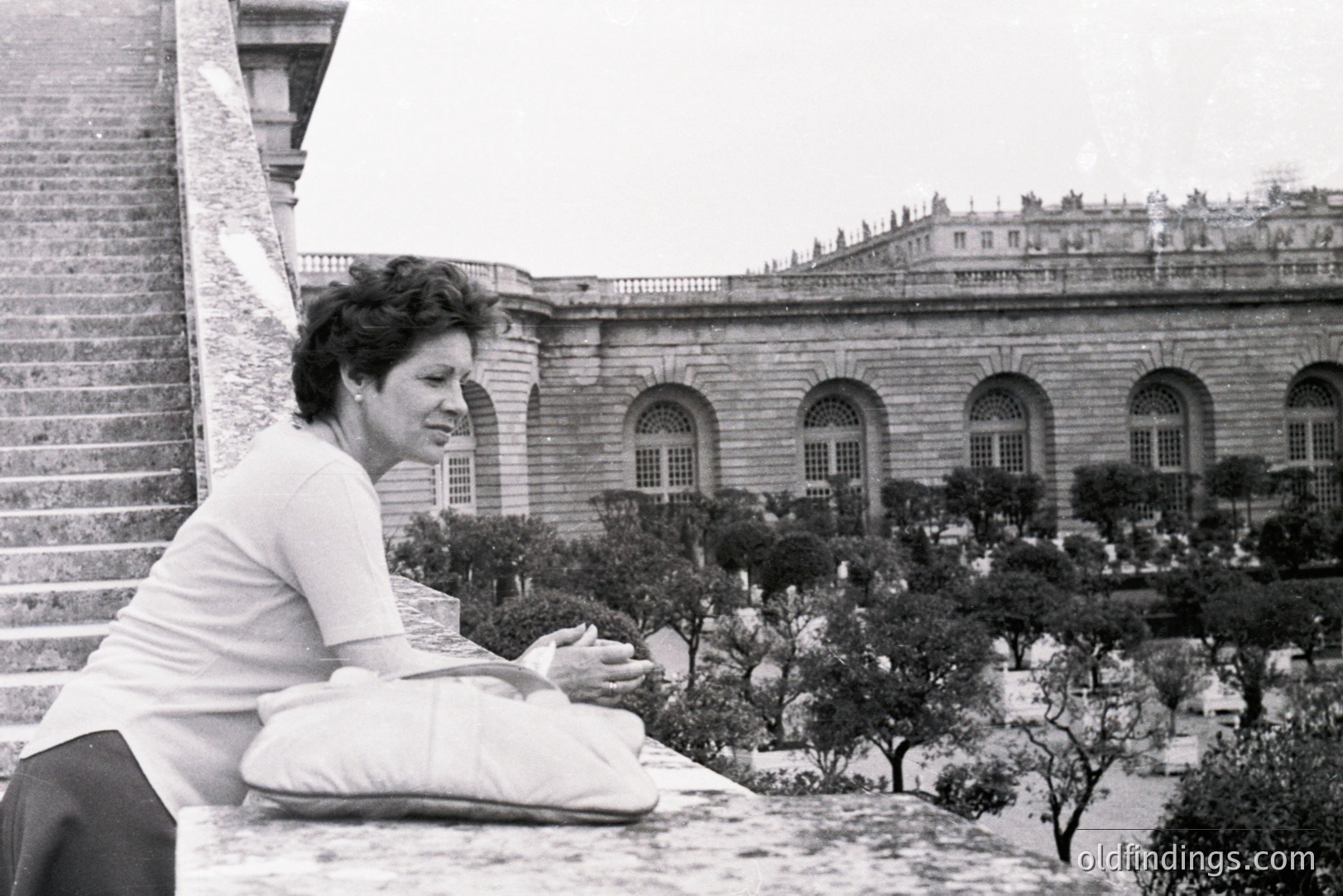 A woman in a simple top and skirt rests on a stone ledge, overlooking formal gardens and a large, ornate building with arched windows. The architectural style suggests a European estate, possibly Italian. Appears to be a candid, mid-century snapshot. Captures a moment of quiet contemplation.