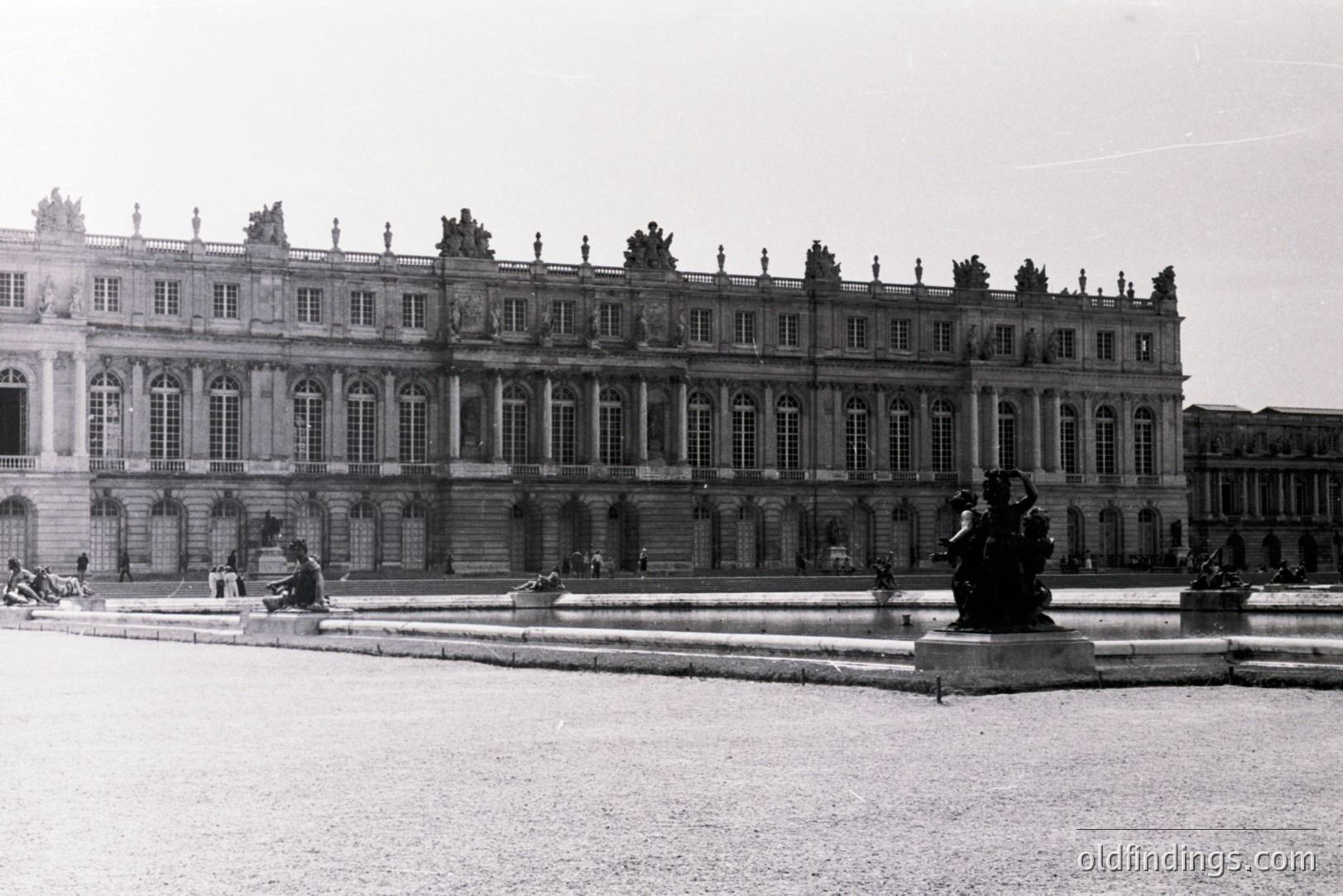 Ornate, symmetrical facade of the Palace of Versailles, France. Classical architecture features numerous windows, elaborate cornices, & a balustrade. A large fountain with sculpture dominates the foreground, surrounded by manicured gardens. Likely captured mid-20th century.