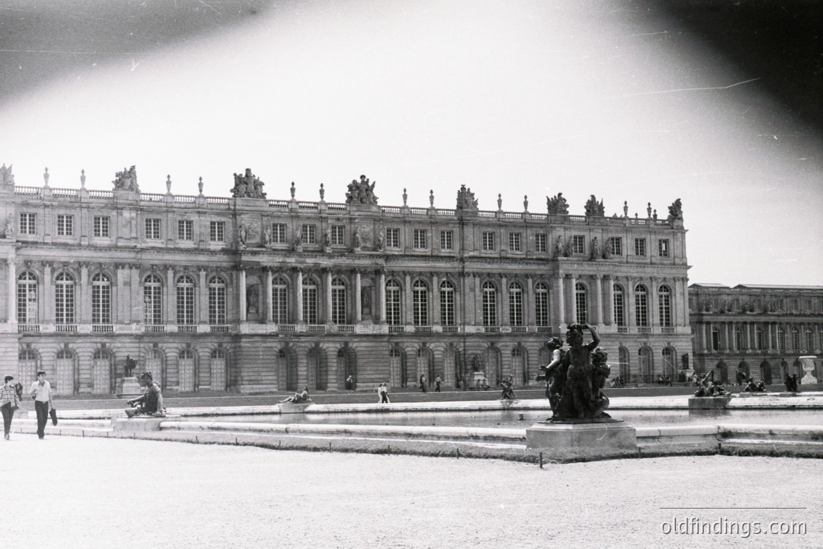 Grand view of the Palace of Versailles, France, showcasing its elaborate Baroque architecture with tiered facades, numerous windows & sculpted details. Formal gardens and statues visible in the foreground. Likely dating from the mid-20th century based on photographic style.