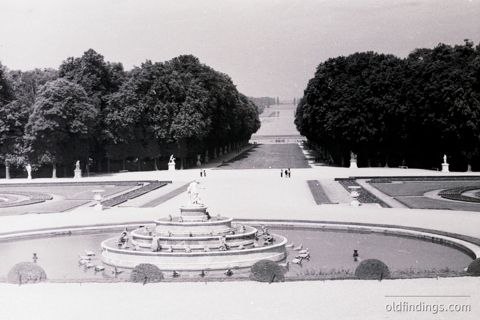Formal French garden with tiered fountain, statues, and meticulously manicured hedges. View extends down a long, straight allée toward a distant structure. Likely a royal or aristocratic estate, mid-20th century. Demonstrates classical garden design principles. Suitable for architectural reference.