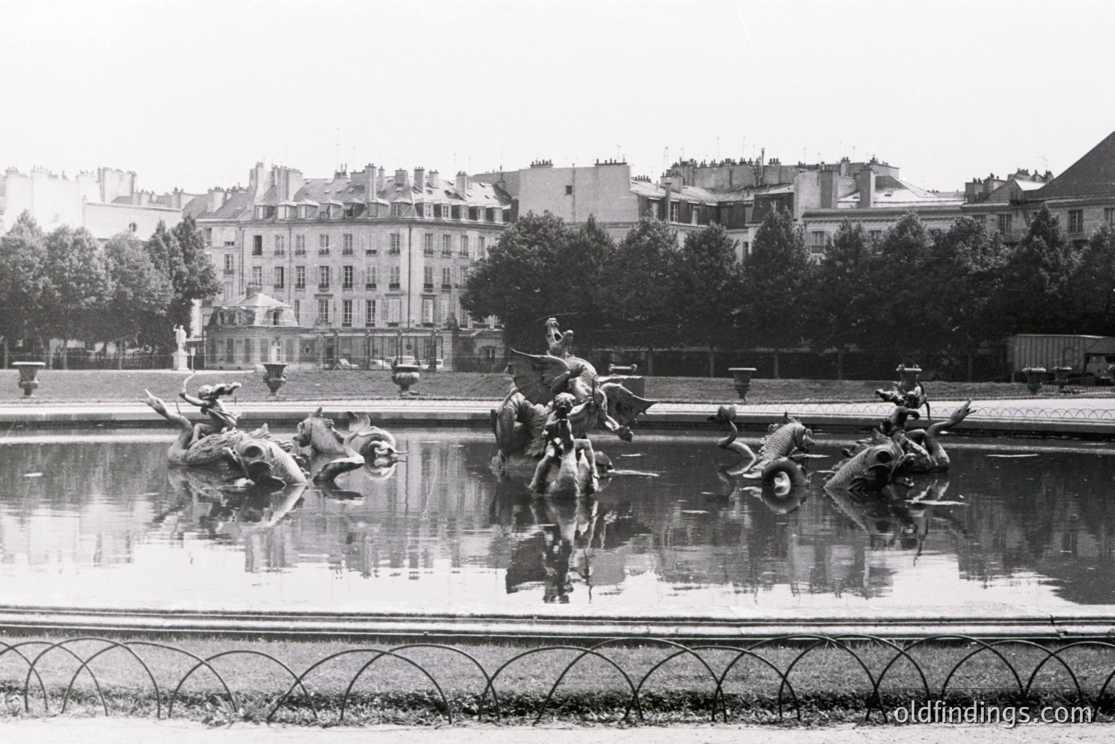 Ornate fountain sculptures reflecting in still water, set within a formal garden. Background reveals Parisian architecture—multi-story buildings with ornate facades. Likely taken in the early-mid 20th century, judging by photographic style. Potential stock imagery reference for landscape or fountain design.