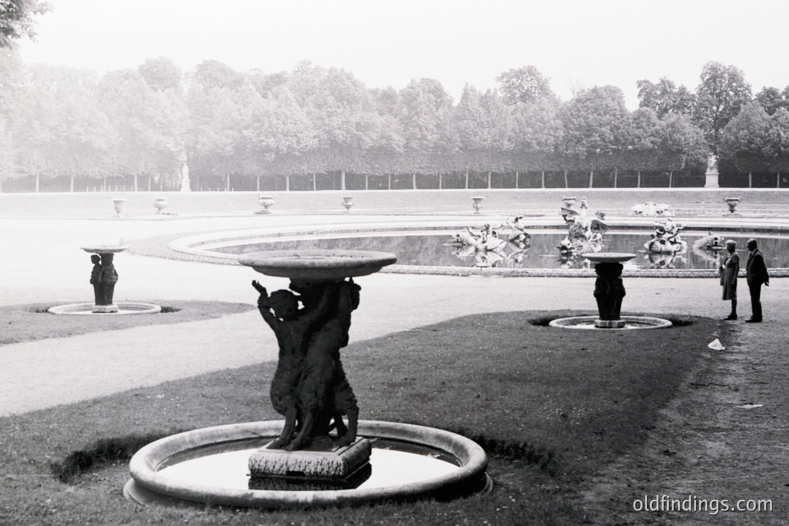 Formal garden scene with tiered fountains & classical statuary. Three fountains visible, central one depicts a figure supporting a stone table. Likely European estate, mid-20th century. The expansive landscape shows meticulous design. People viewed from a distance. garden
