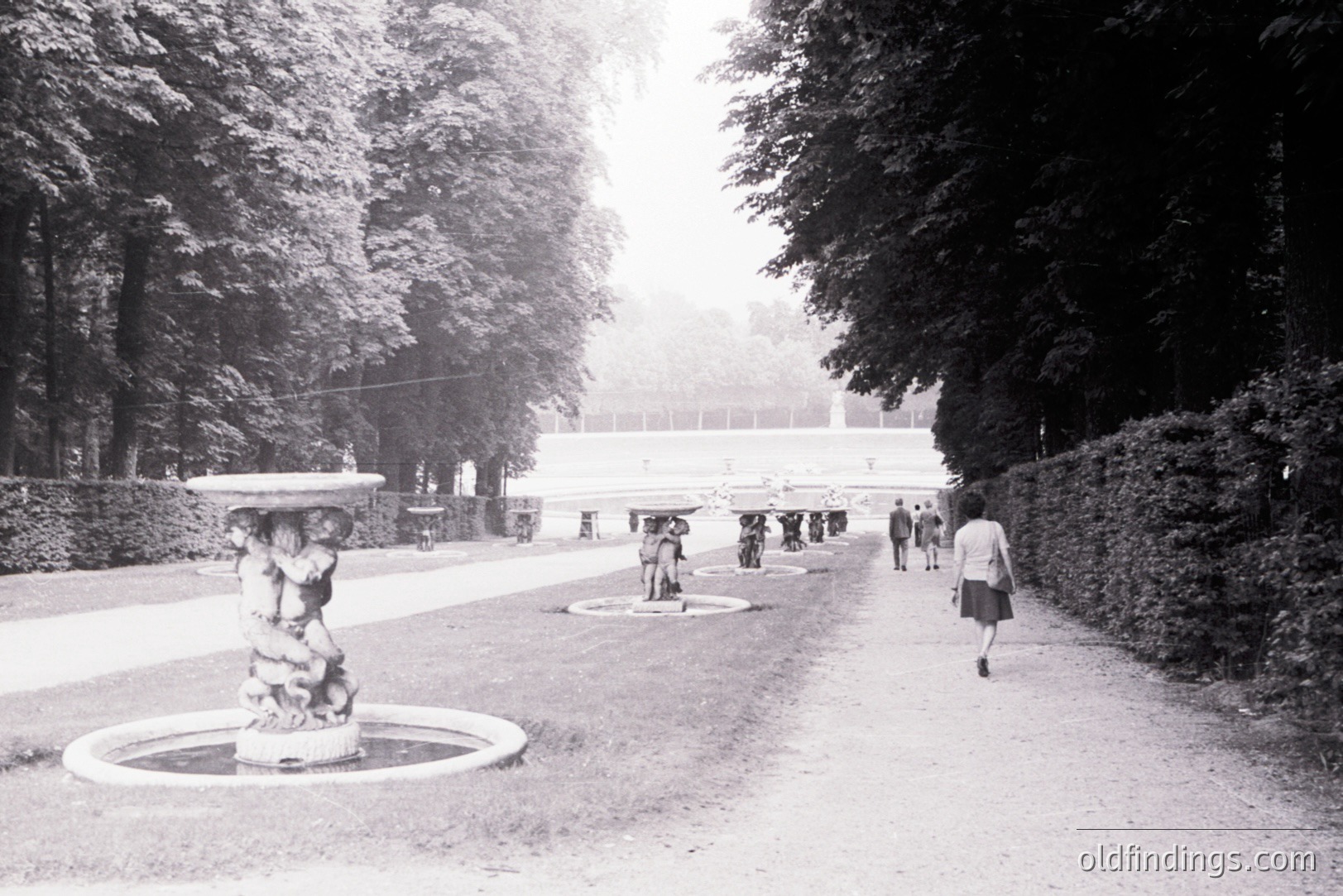 Ornate fountain and gravel path lead towards a formal garden facade. Several figures are visible in the distance, strolling amongst tiered landscaping. Likely a European estate, possibly 1960s or 70s. Evokes a sense of stately elegance and quiet leisure. garden
