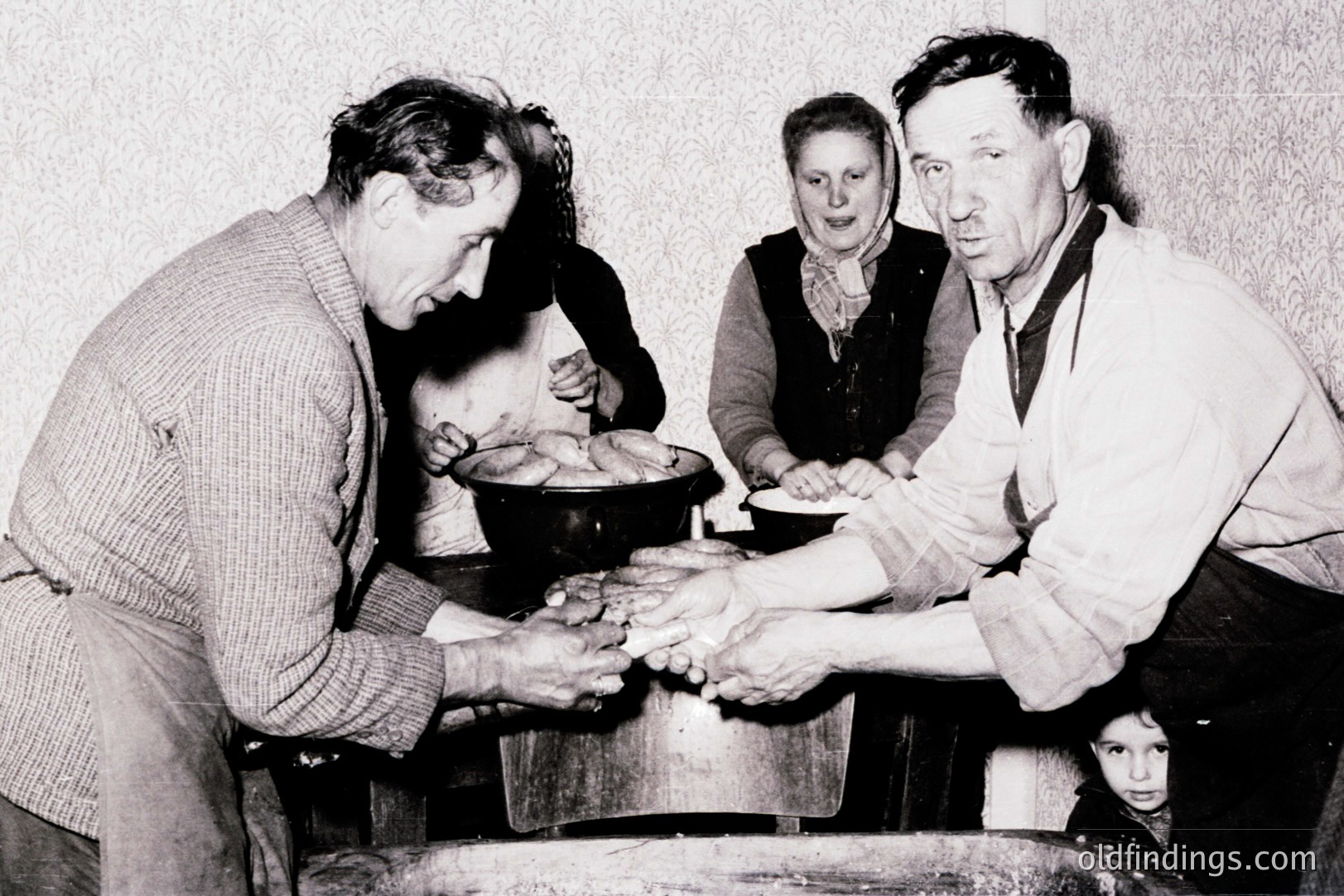 A family gathers around a large pot, seemingly making or preparing cheese. A man kneads dough while another gestures, possibly instructing. A young girl peeks from behind. Simple, domestic setting; likely rural or working-class household. Estimate: 1950s-1970s, Eastern Europe. Captures a moment of traditional food preparation.