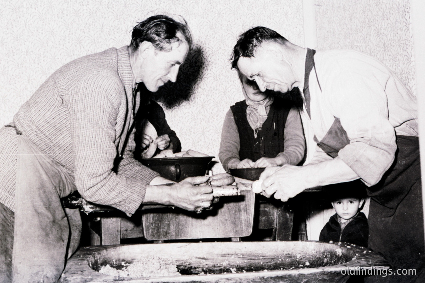 Two men work together at a traditional mill, likely processing grain. A young boy observes from the shadows. The scene suggests a rural, agricultural setting, possibly Eastern Europe. The attire and mill design indicate a mid-20th century timeframe (1940s-1960s). A valuable documentation of traditional practices.