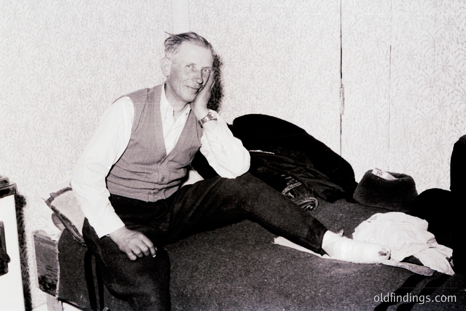 Man seated on a vintage sofa, showcasing a leg encased in a cast. He wears a waistcoat over a collared shirt and dark trousers. A hat and bag rest nearby, suggesting travel. Likely a candid portrait from the 1940s-1960s.