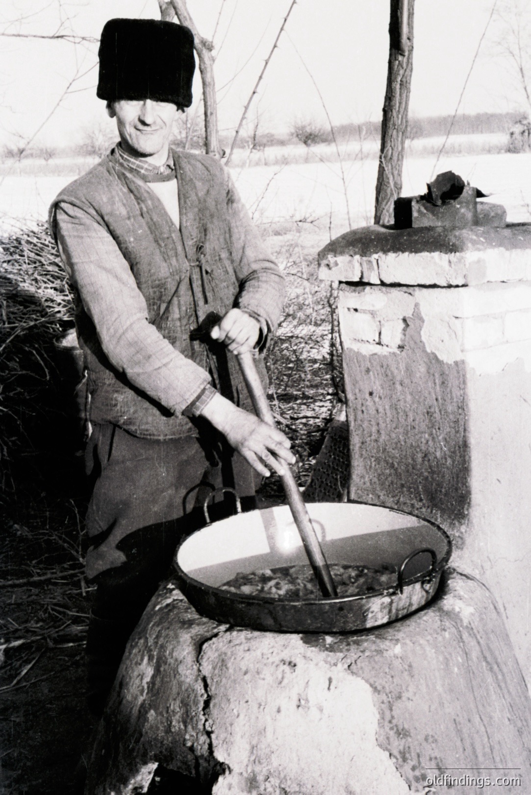 A man wearing a fur hat and dark clothing vigorously stirs a large cauldron set on a stone platform, likely involved in food or beverage production. Outdoor setting with a brick structure visible. Likely a rural scene depicting traditional processing methods. Estimated timeframe: mid-20th century. Potential for historical foodways documentation.