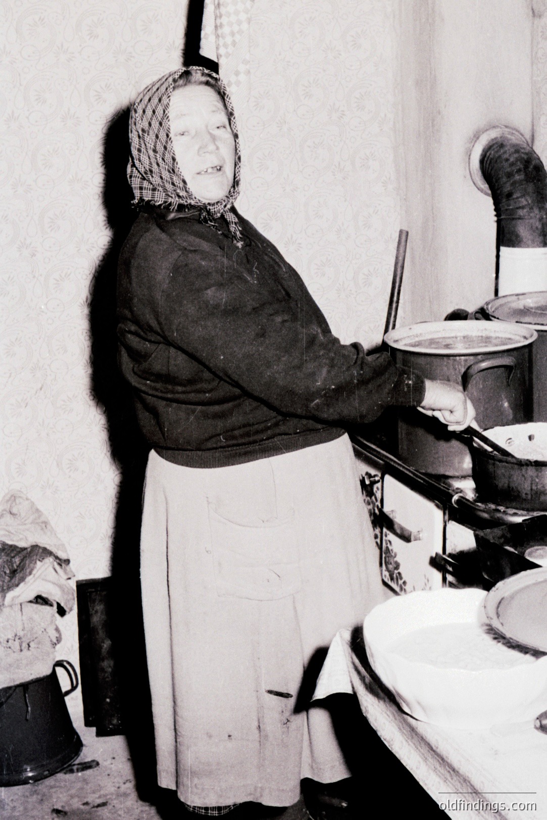 A woman in traditional clothing tends to a large stove in a rustic kitchen. She wears a dark dress, white apron, and headscarf. The stove features a prominent chimney and multiple cooking pots. Likely domestic scene, potentially documenting rural life or food preparation.