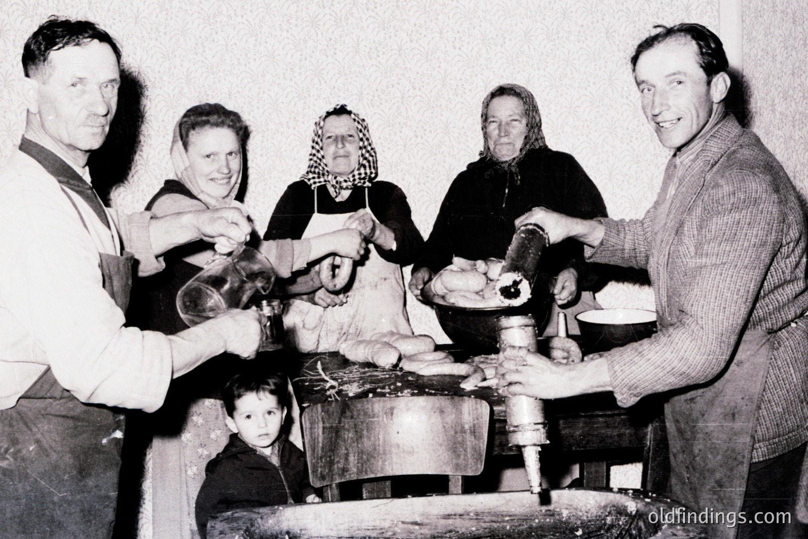 A family workshop scene, likely cheese or dairy production. Five figures are shown – a man in an apron, two women in headscarves, another man, and a child. Equipment includes a large bowl, churn, and tools for processing dairy. Likely rural, Eastern European, mid-20th century.