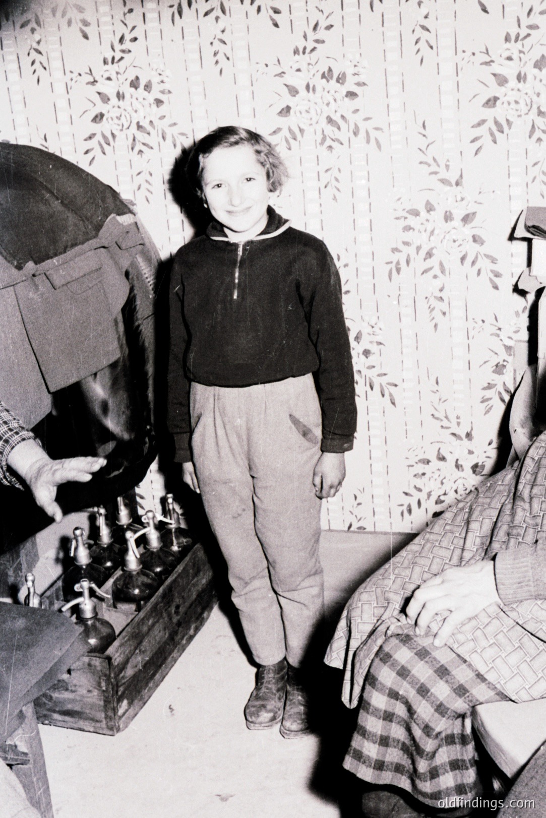 A young boy stands before a decorative display of bottles, likely a pharmacy or shop interior. He wears a zippered sweater and corduroy trousers. The wallpaper & visible furnishings suggest a domestic setting, possibly in the 1940s. A woman is partially visible in the background.