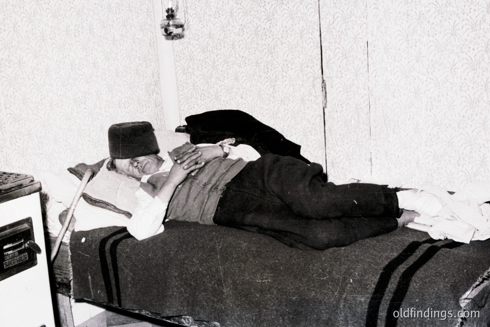 Man lies on a narrow bed, propped up by pillows, wearing a traditional hat and vest. A medical device sits beside the bed in a sparsely furnished room. Likely a portrait documenting medical treatment, possibly tuberculosis sanatorium. Estimated: 1930s-1950s.