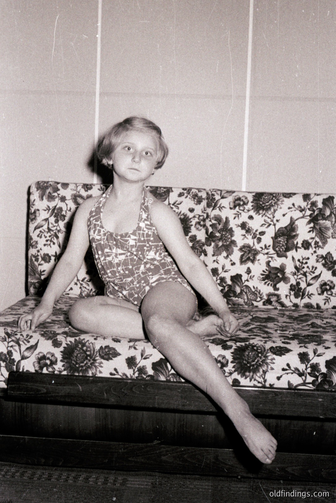 Young girl in a one-piece bathing suit, seated on a floral-patterned bench or sofa. Posed portrait, likely mid-20th century, possibly 1950s or 1960s, based on hairstyle & photographic style. Interior setting, neutral-toned background. Potential for vintage advertising/lifestyle reference.