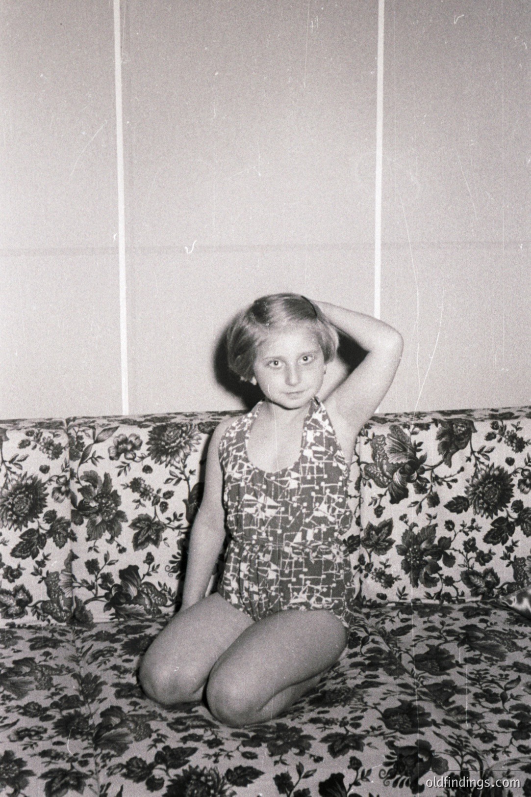 A young girl in a patterned one-piece swimsuit sits cross-legged on a floral-print sofa. She's touching her hair, facing the camera with a neutral expression. The background appears to be a plain wall. Likely a 1960s lifestyle or family portrait. Potential design reference for vintage textiles.