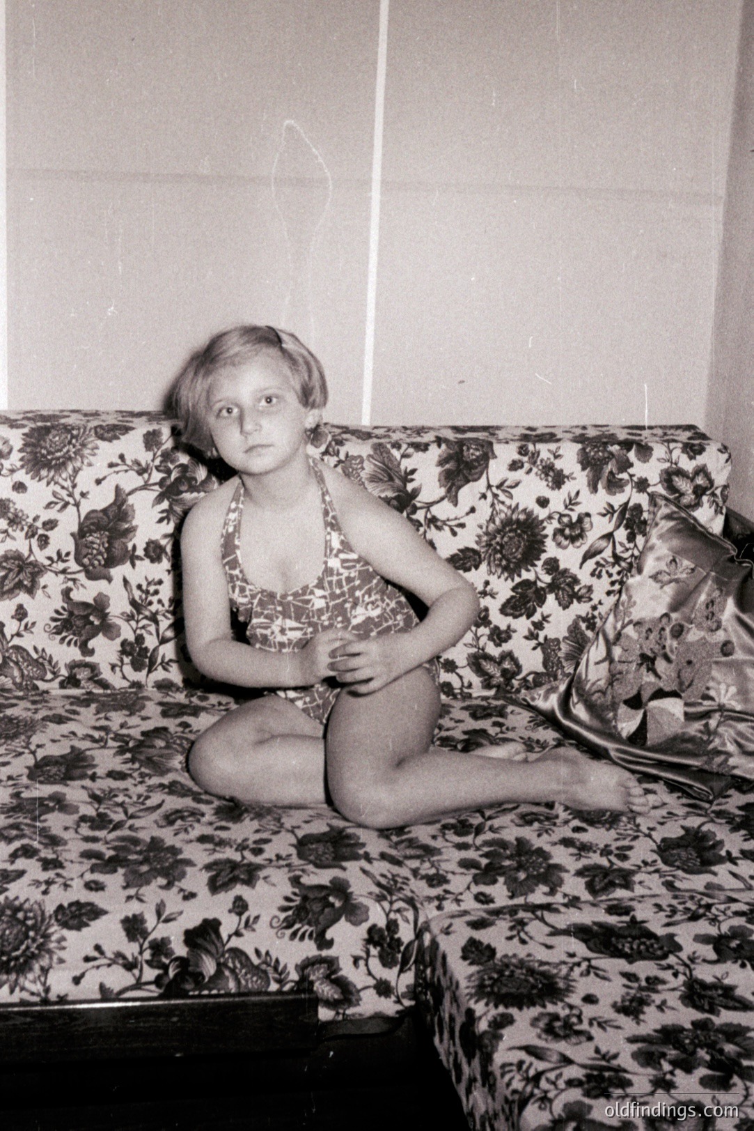 Young girl in a patterned swimsuit sits on a floral-patterned sectional sofa. Interior scene, likely a holiday accommodation. Possible 1960s-1970s era; the style of the swimsuit and furniture suggest this timeframe. High commercial value for design reference and nostalgia.