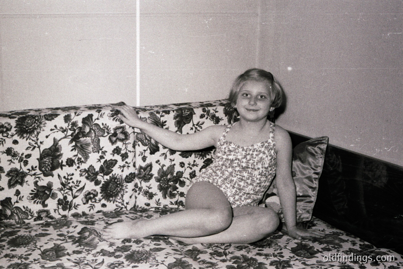 A young girl, circa 1960s-70s, sits cross-legged on a floral-patterned couch, wearing a one-piece bathing suit. Her arm is extended, casually posed. Black and white photo; likely a personal snapshot or studio portrait. Minimal background. Commercial value: design reference, nostalgic portraiture.