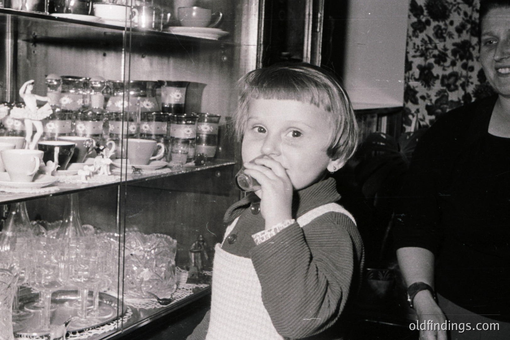 A young child stands before a glass display case filled with delicate china and figurines, appearing to savor a treat. The child wears a striped sweater and apron. A partial view of an adult is visible in the background. Likely a shop interior, mid-20th century.