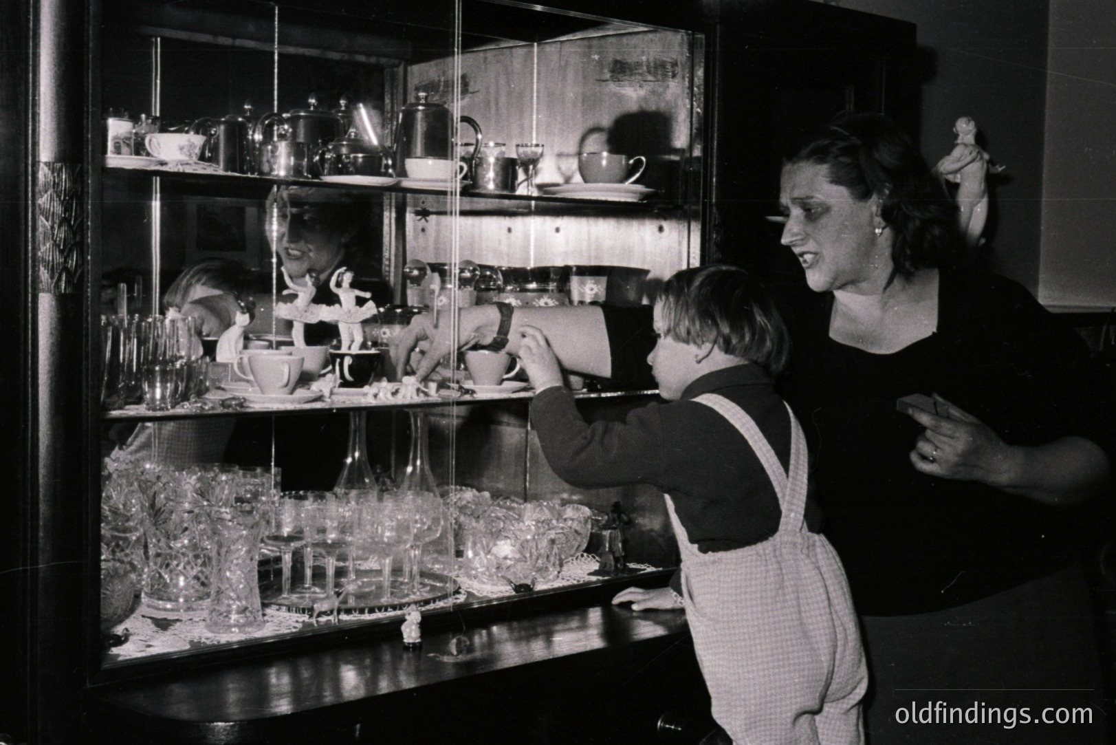 Young boy in overalls examines a glass cabinet filled with china, silver, and figurines. A woman stands behind him, her expression curious. Likely a domestic interior, possibly 1950s. Captures childhood curiosity & mid-century design aesthetics. Potential for stock or vintage design references.