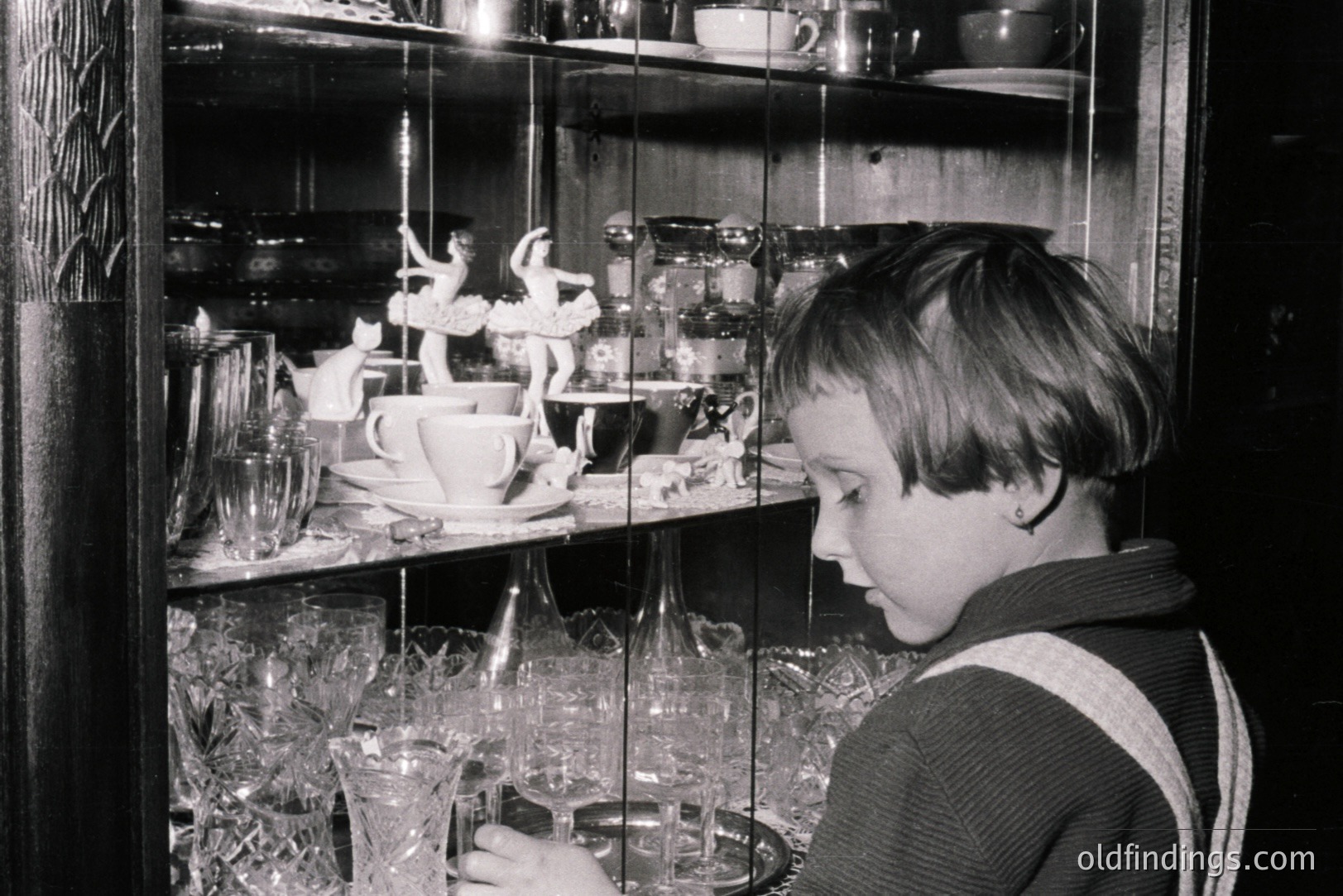Young boy intently examines a glass display cabinet filled with ornate figurines, glassware, and tableware. Likely a domestic interior, the scene evokes a sense of quiet observation and curiosity. Style suggests mid-20th century (1950s-1970s). Valuable as a representation of childhood and home life.