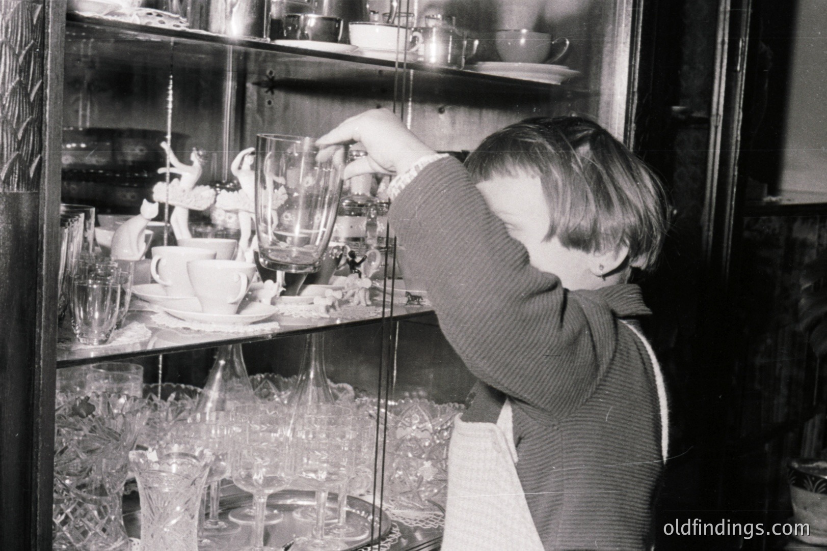 A young boy reaches for a glass goblet within a crowded china cabinet. The cabinet displays delicate glassware, ornate figurines, and patterned china. The scene evokes a domestic, possibly formal, setting. Likely a candid snapshot from the mid-20th century.